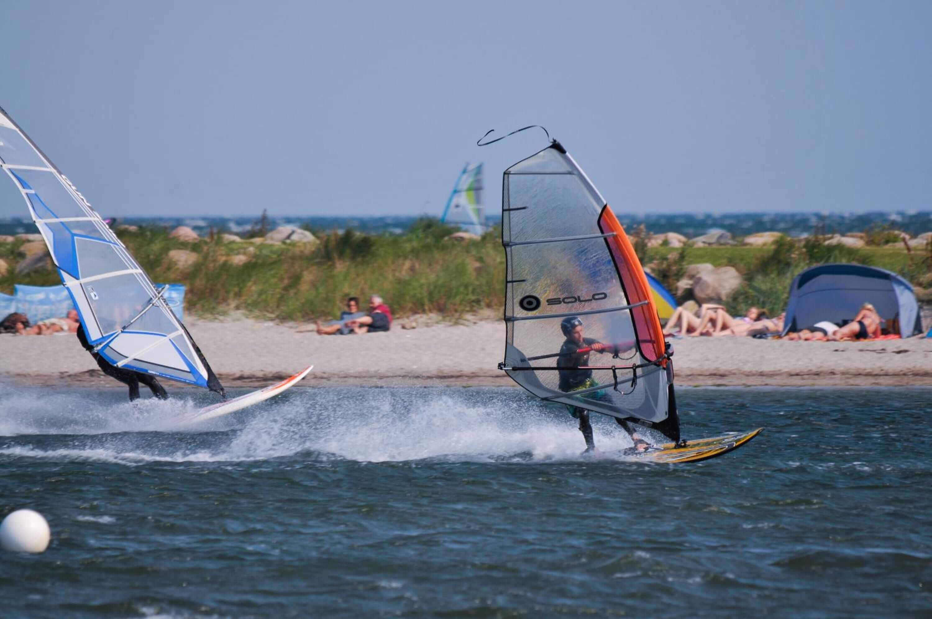 Zwei Windsurfer am Strand, im Hintergrund Zelte und Personen.