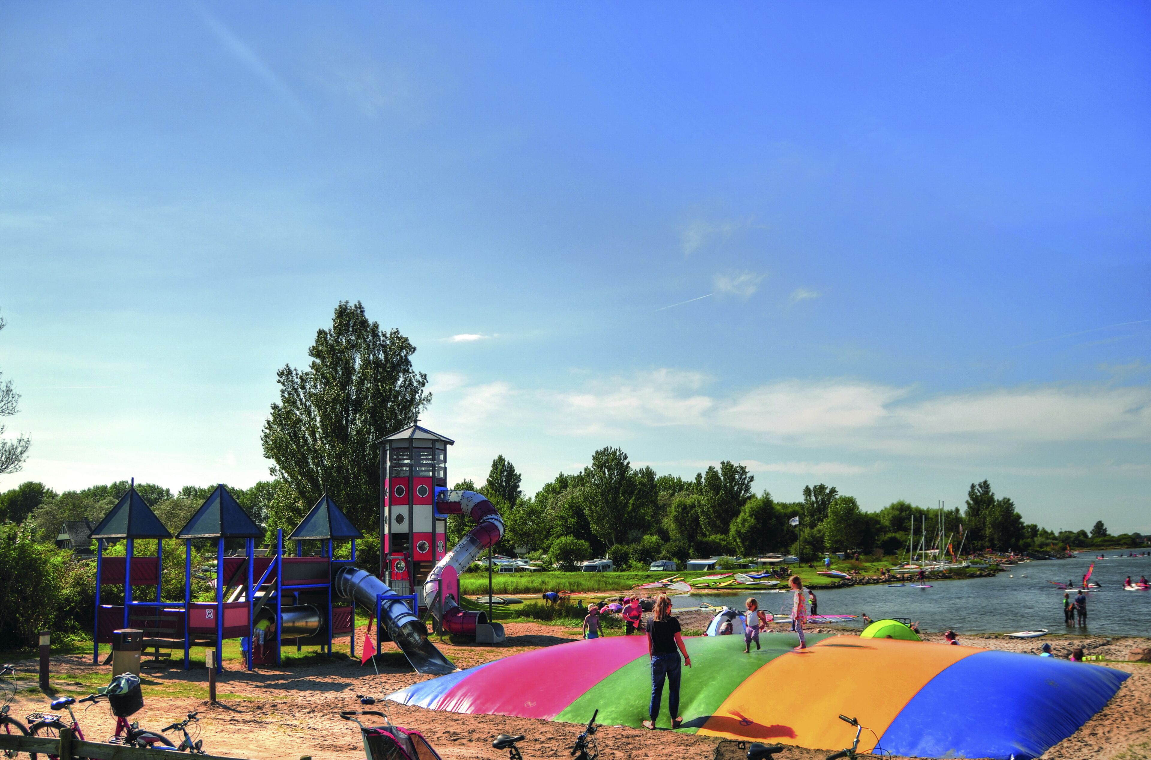 Un parque infantil con toboganes y estructuras para escalar, una zona de playa con arena y un río con barcos.