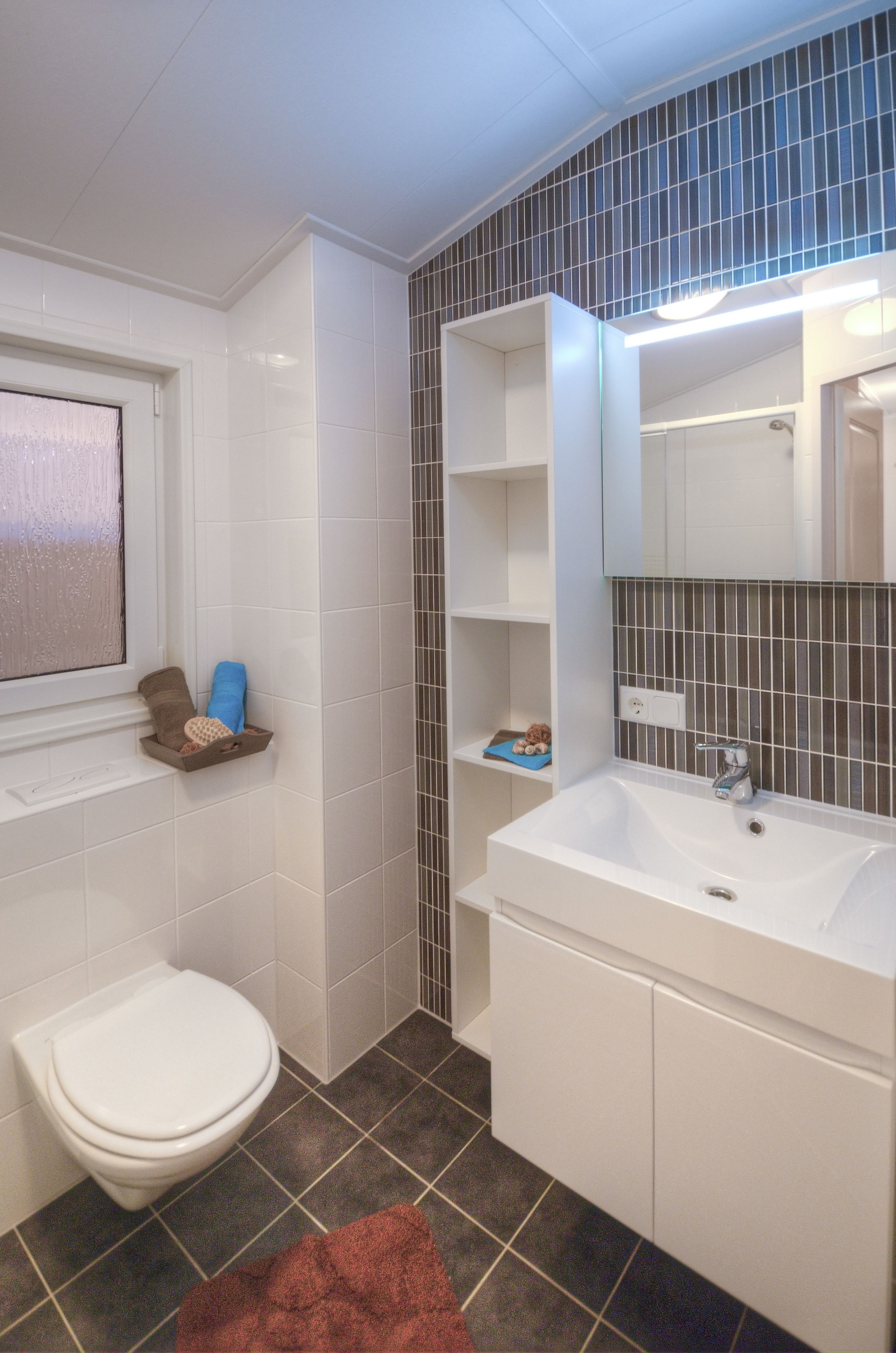 Une salle de bain avec des toilettes blanches, un lavabo avec un miroir et des étagères remplies d’articles de toilette.