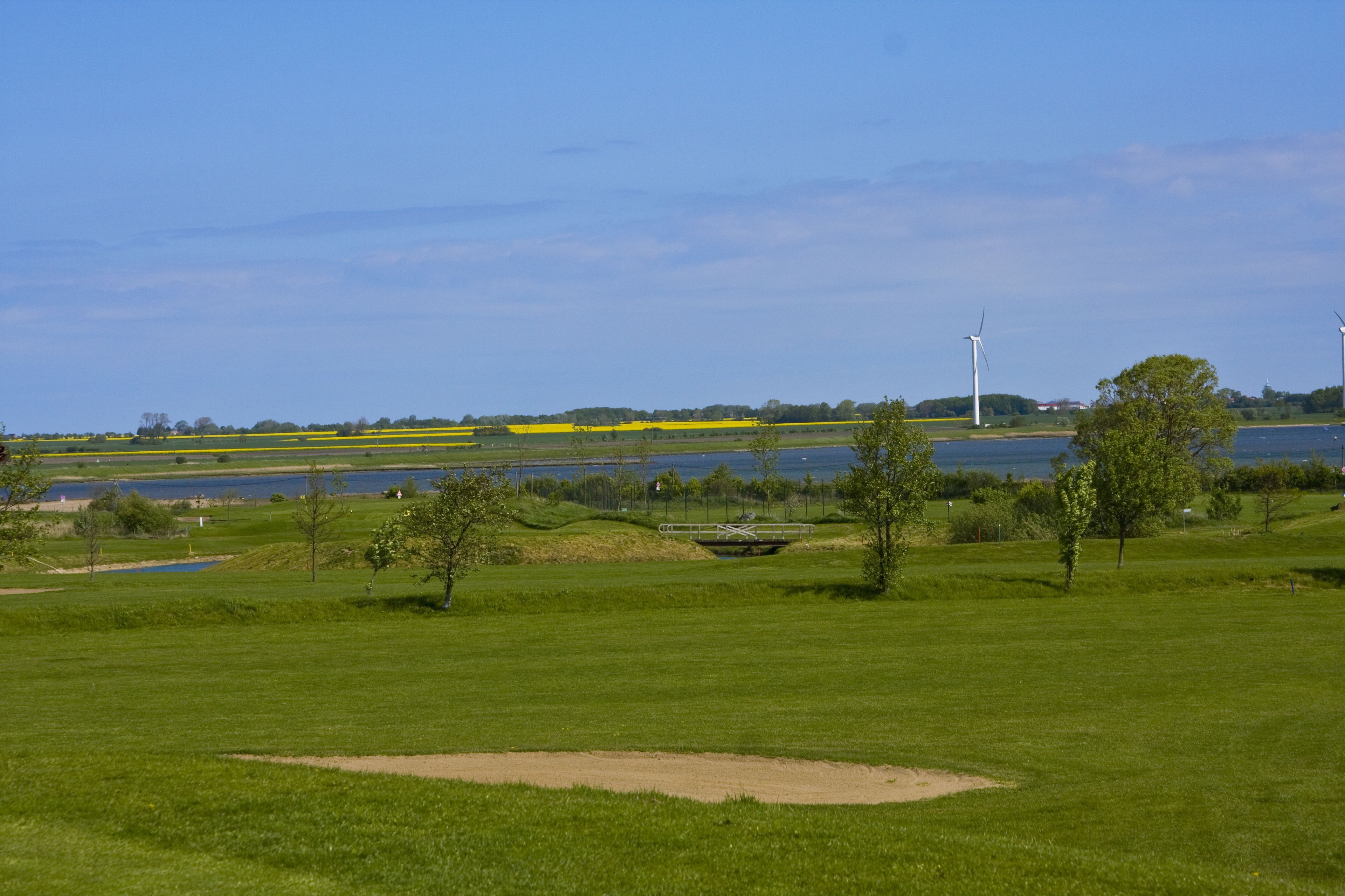 A golf course with a water body and wind turbines in the background.