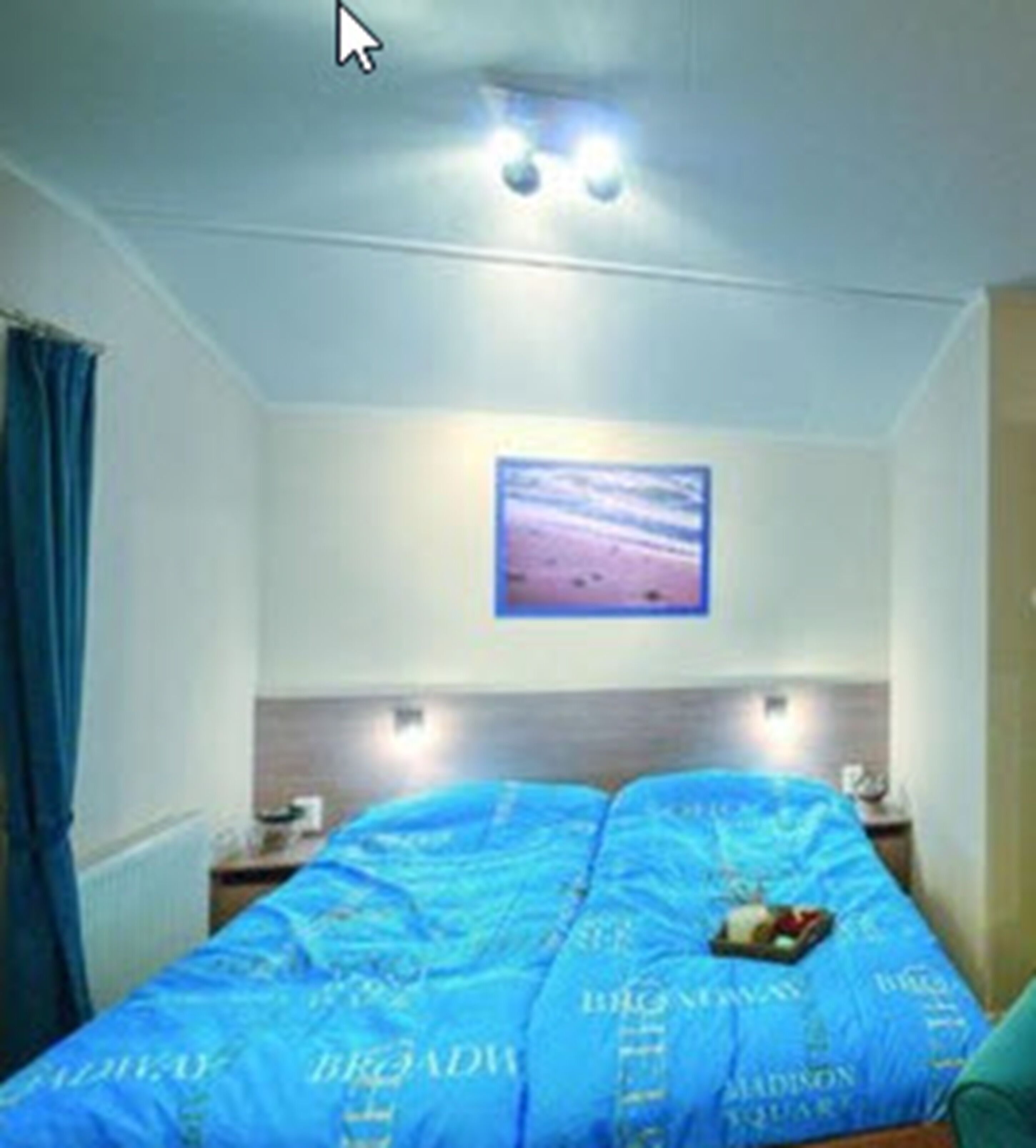 Une chambre avec un lit recouvert d’une literie bleue, une table de chevet avec une lampe et un tableau encadré au mur.