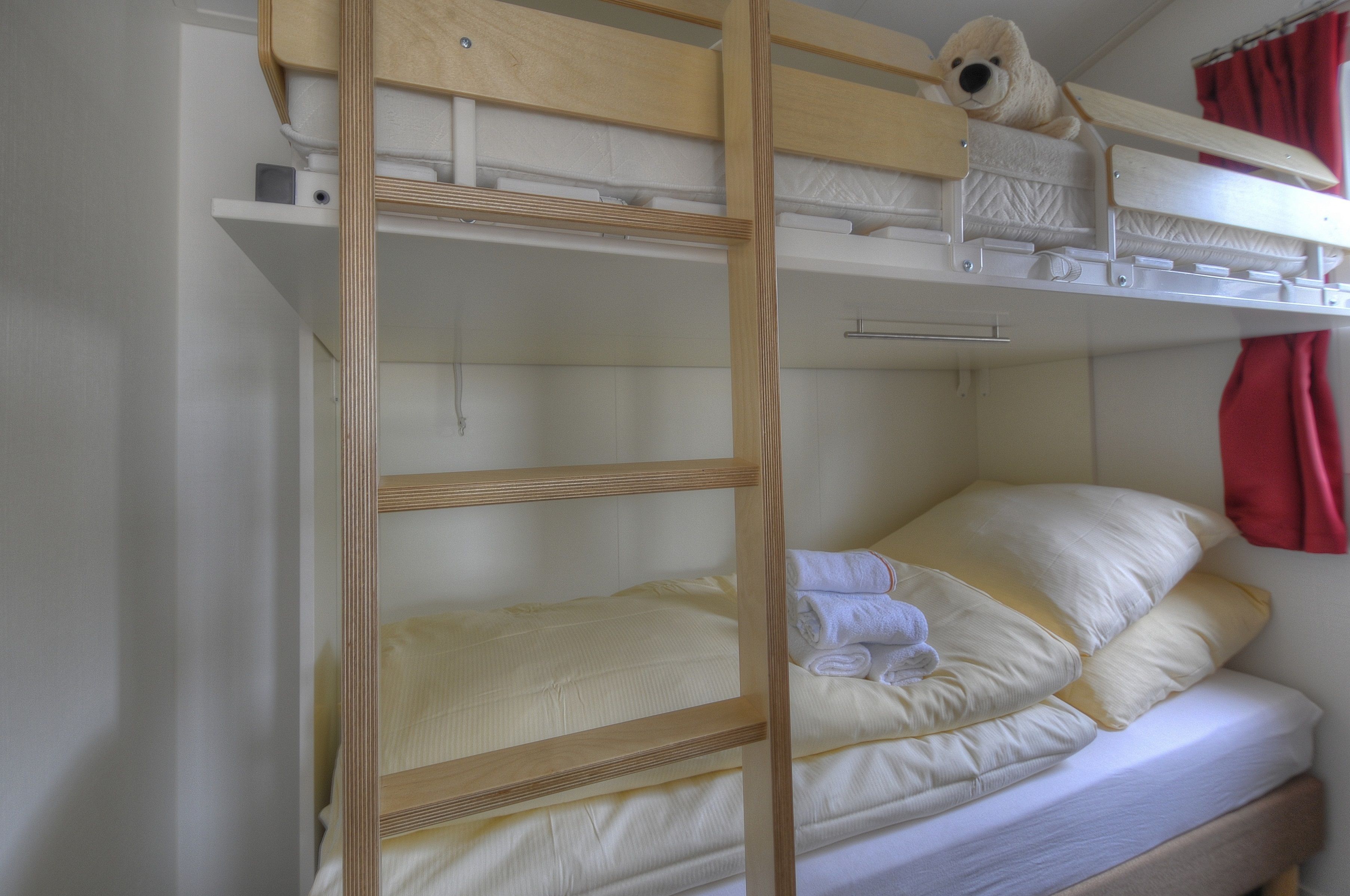 A bunk bed with a wooden ladder, a teddy bear on the top bunk, and a red curtain in the background.