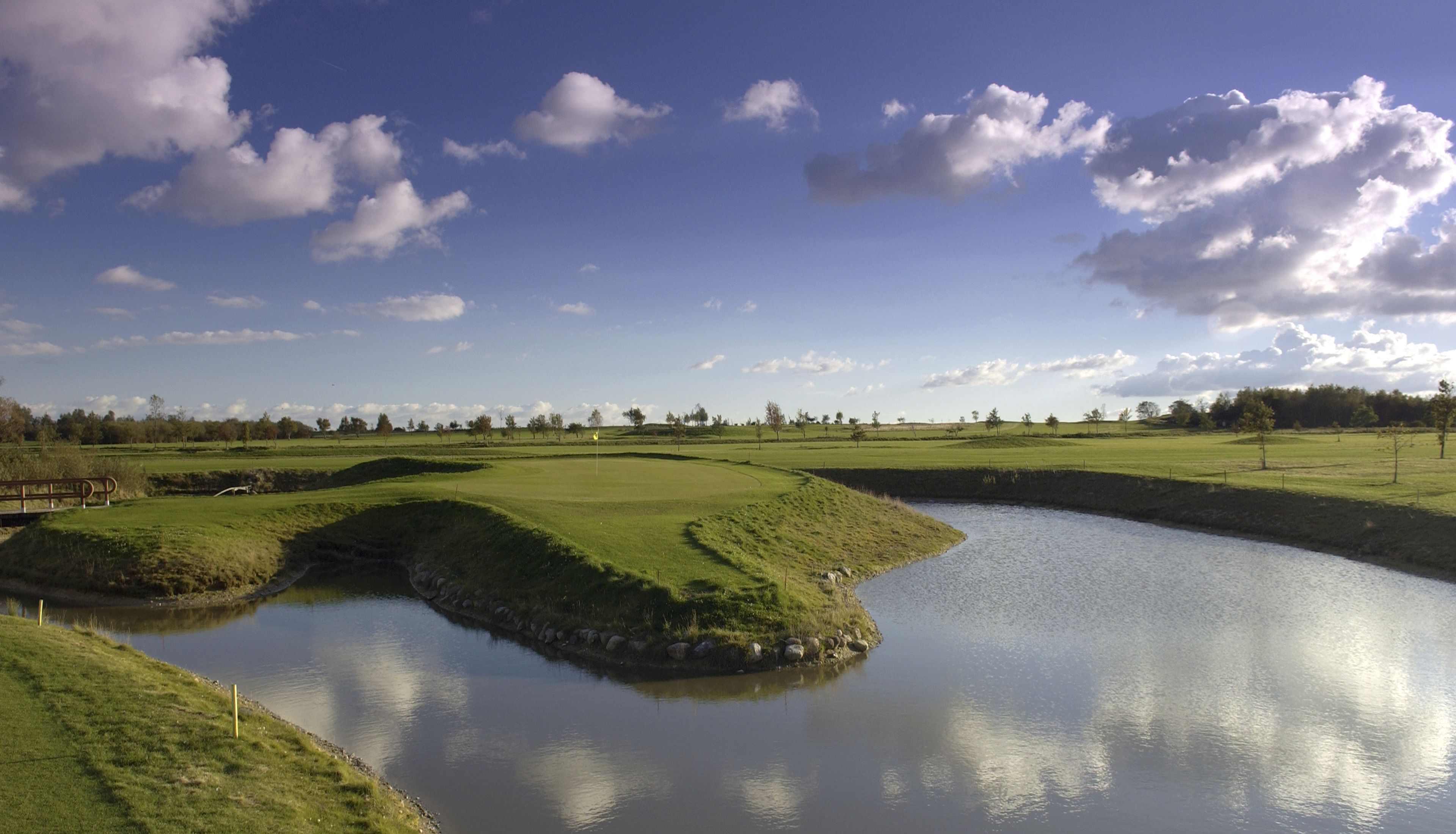 Un campo de golf con un estanque, rodeado de campos verdes y árboles bajo un cielo parcialmente nublado.