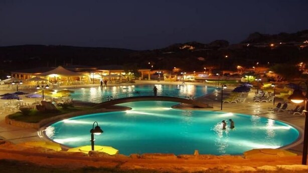 Vista nocturna de una piscina con un puente, rodeada de zonas de descanso y mobiliario exterior.