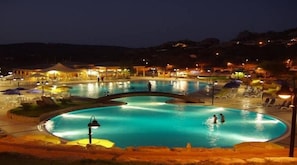 A nighttime view of a swimming pool with a bridge, surrounded by lounge areas and outdoor seating. - Rena Majore Resort (Aglientu)