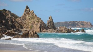 Una costa rocosa con olas rompiendo contra la orilla y grandes formaciones rocosas al fondo.