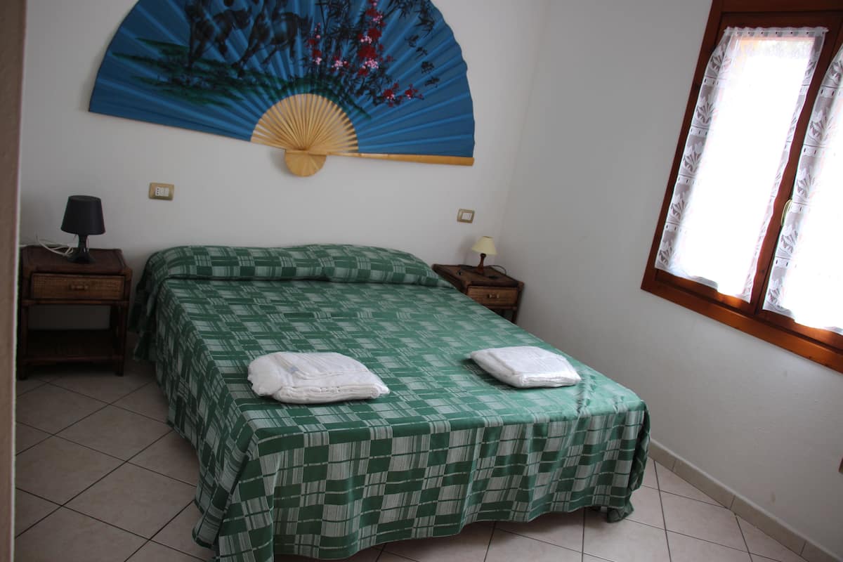 A room with a bed, two bedside tables, a wall-mounted fan, and a window with curtains.