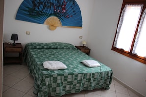 A room with a bed, two bedside tables, a wall-mounted fan, and a window with curtains. - Rena Majore Resort (Aglientu)