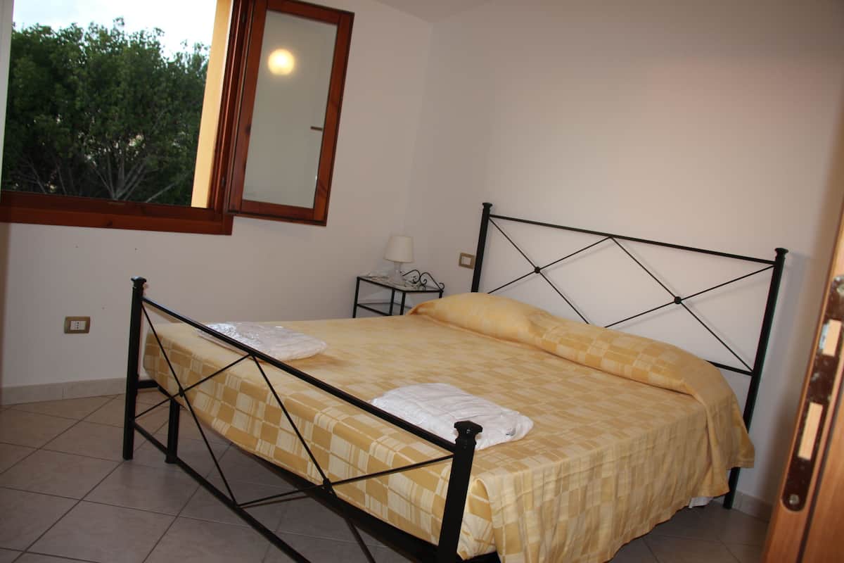 A single bed with a metal frame, a yellow checkered bedspread, and two white towels. There is a small bedside table with a lamp, a window with a view of trees, and a mirror on the wall.