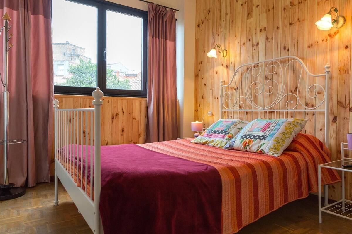 A bedroom with a wooden paneled wall, a white metal bed frame, a red bedspread, and two pillows with floral patterns.