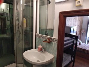 A bathroom with a glass shower enclosure, a sink with a soap dispenser, and a bed with a striped bedspread.