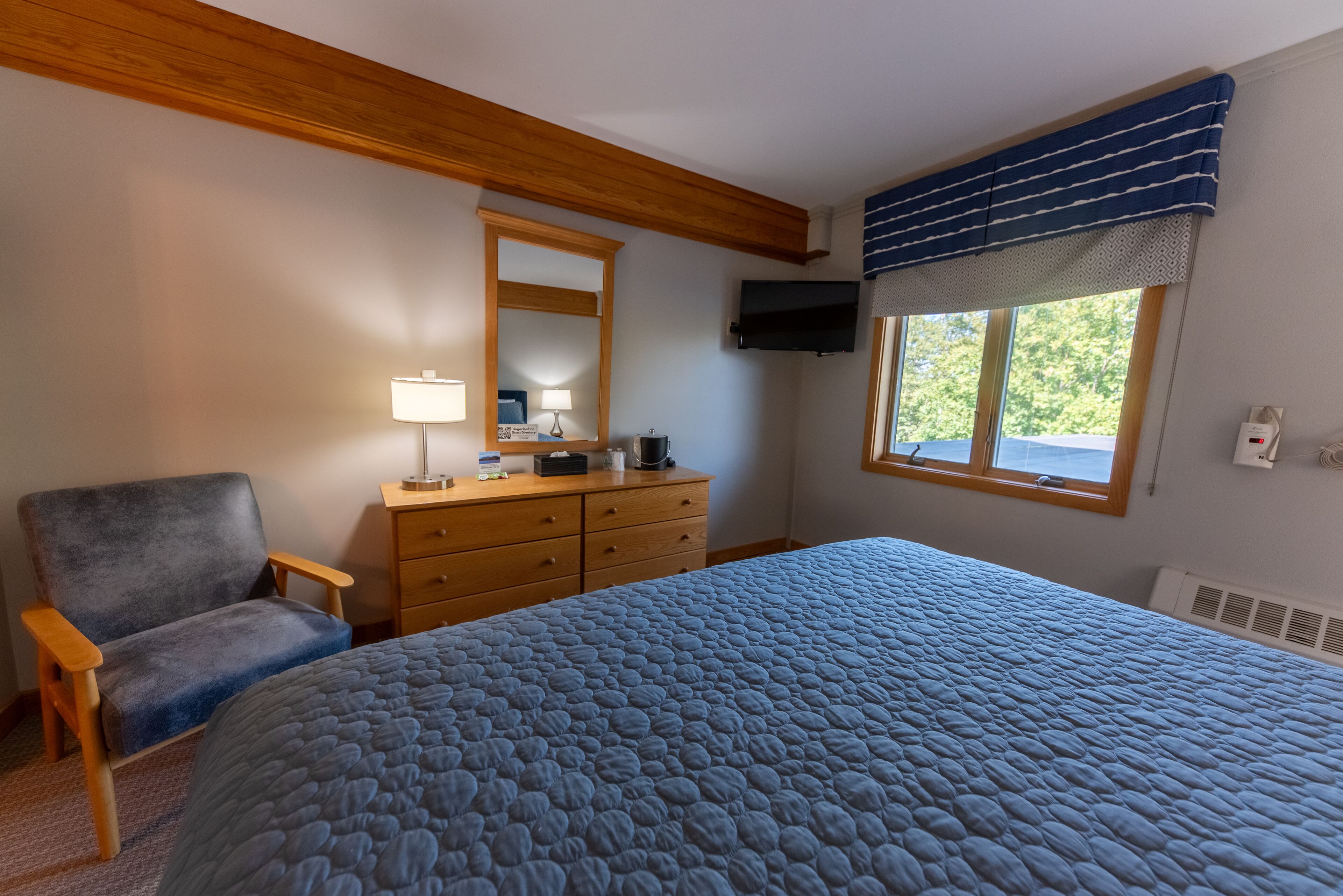 A bedroom with a large bed, a wooden dresser, a chair, a TV mounted on the wall, and a window with a view of trees.