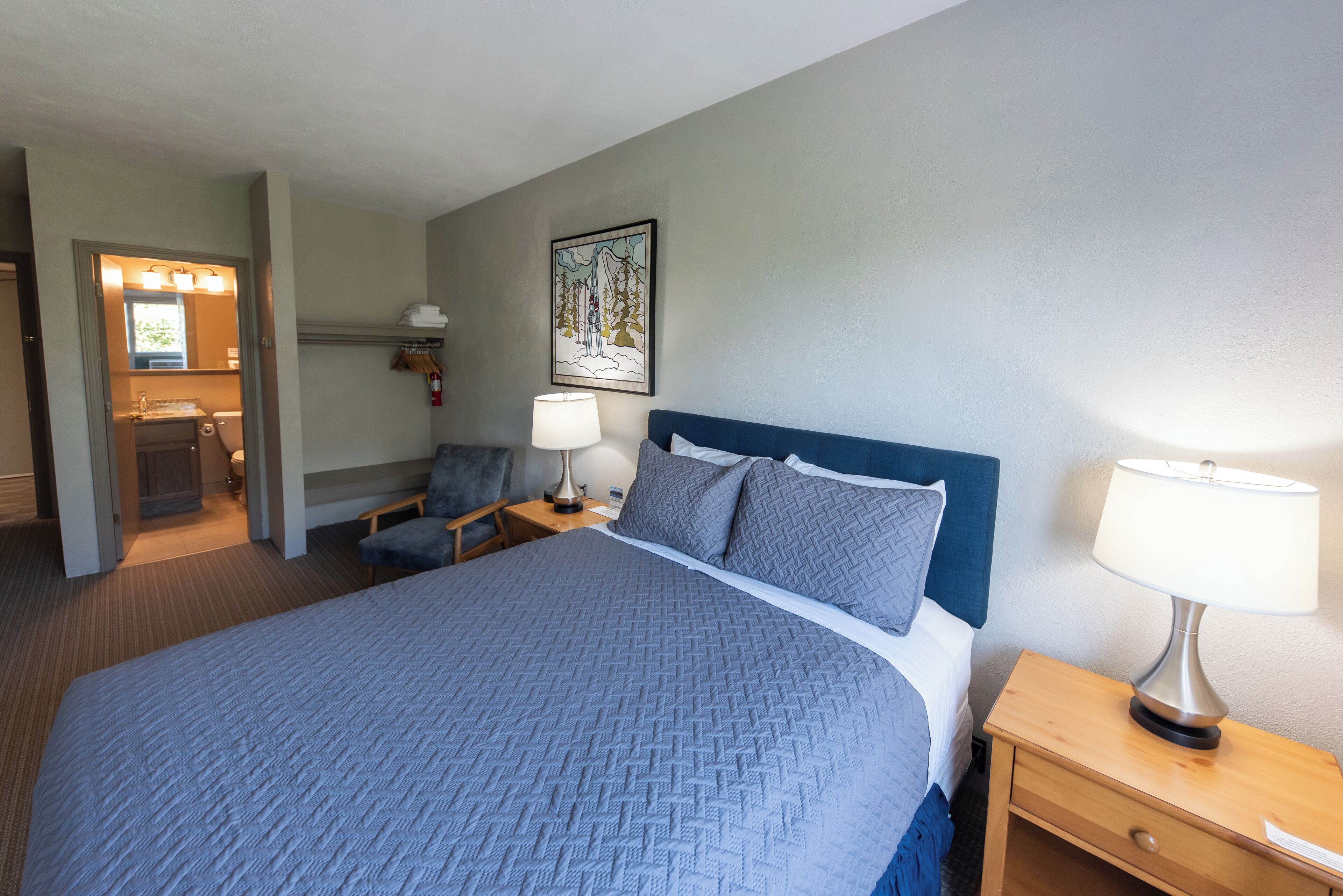 A hotel room with a large bed, a bedside table with a lamp, a chair, a bathroom with a mirror, and a wall-mounted shelf.