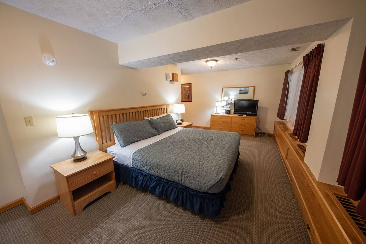 Una habitación de hotel con cama, mesita de noche, lámpara, televisor, cómoda y ventana con cortinas.