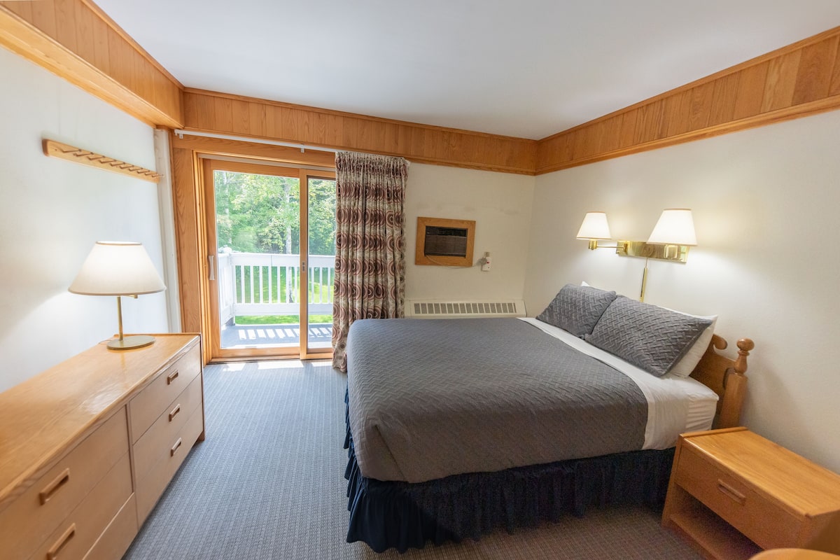 Un dormitorio con cama, mesitas de noche de madera, cómoda, ventana con cortinas y vista a un balcón.