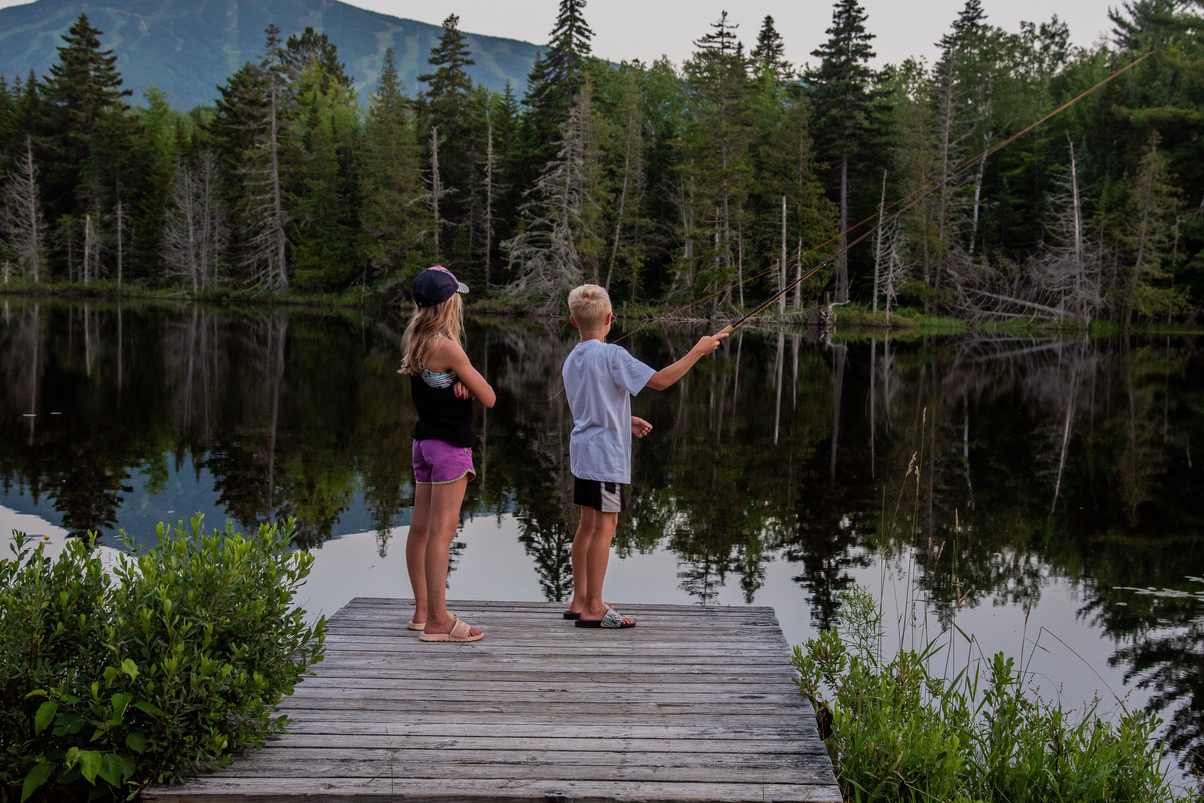 Two people standing on a wooden dock by a calm lake surrounded by dense forest.
