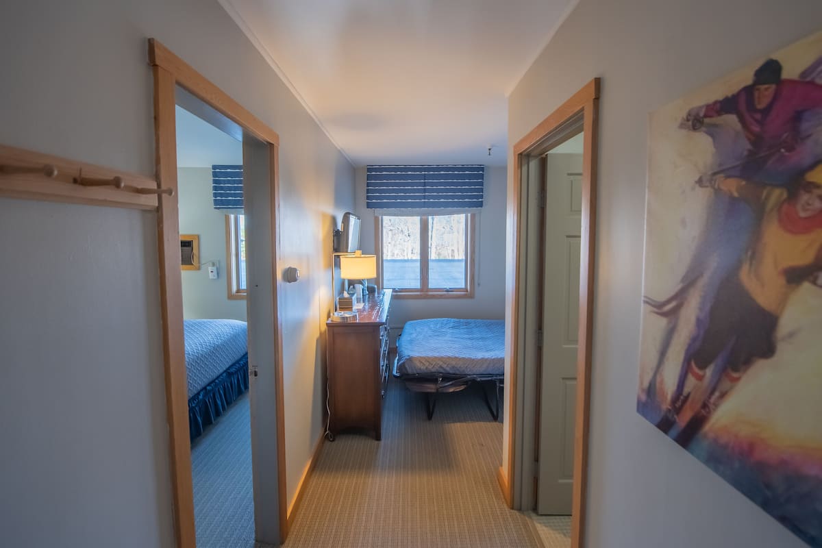 Un pasillo estrecho que conduce a un dormitorio con cama, cómoda y ventana con persianas.
