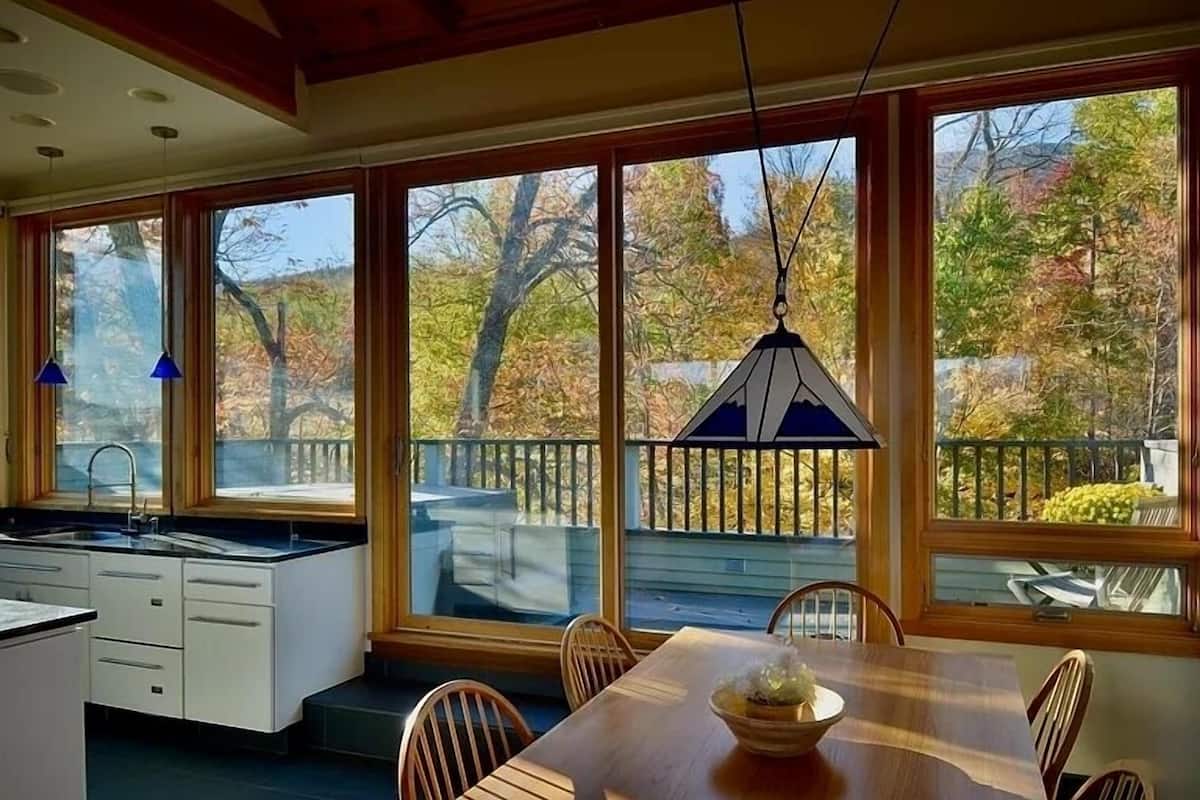 Una cocina moderna con una isla grande, un comedor con mesa y sillas de madera, y ventanas amplias con vistas a árboles y un balcón.