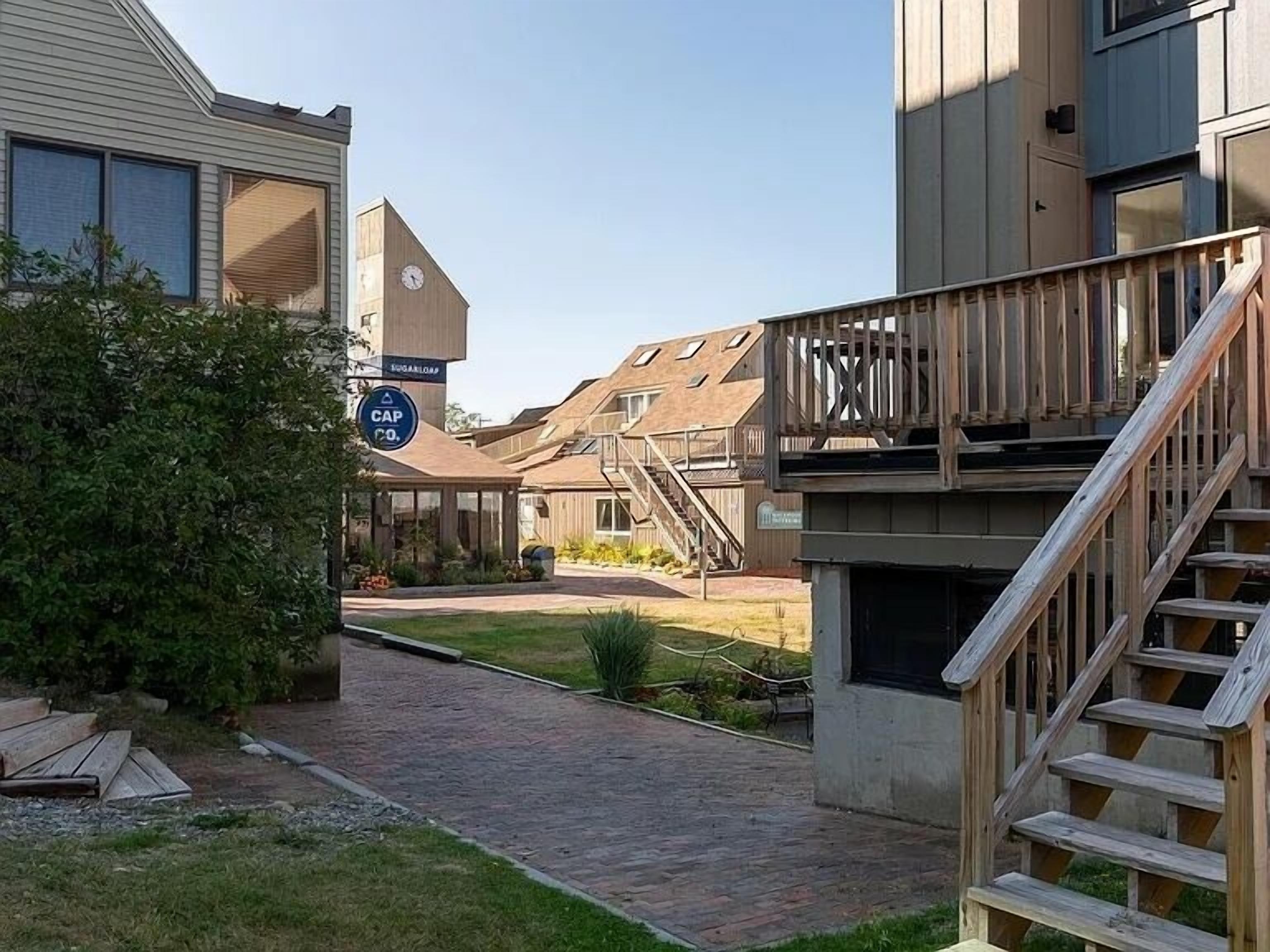 Un quartier résidentiel avec un escalier en bois menant à un balcon, un chemin en briques et un panneau indiquant une entreprise nommée « CAP CO. ».