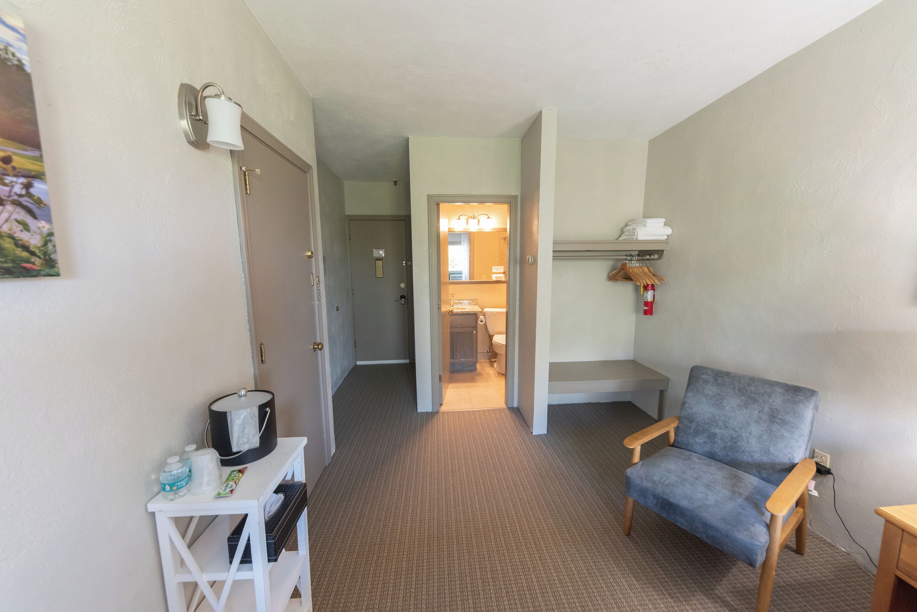 Habitación de hotel con kitchenette, una silla, una mesita y un baño visible a través de una puerta abierta.