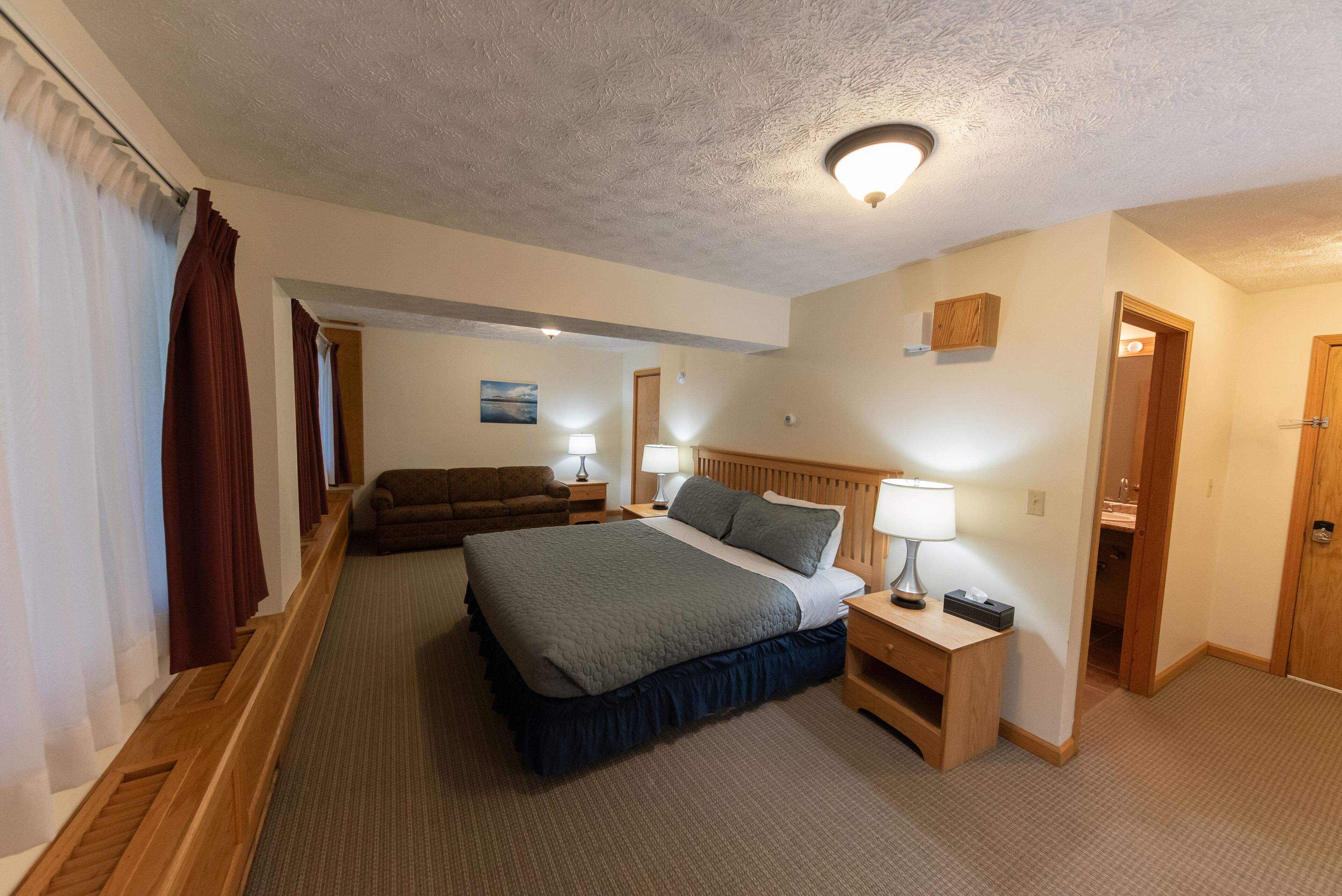 Habitación de hotel con una cama grande, una mesita de noche de madera, un escritorio, una silla y un baño visible a través de una puerta abierta.