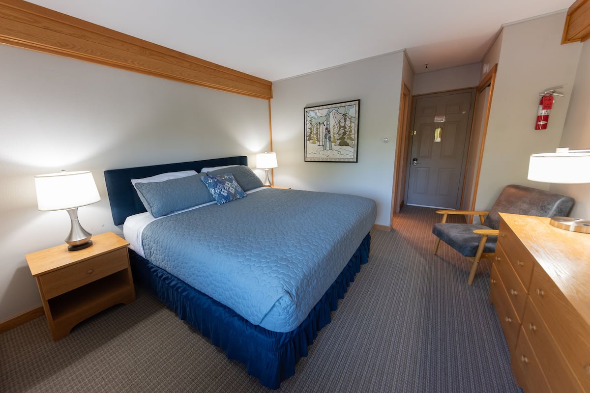 Una habitación de hotel con una cama grande, cabecero de madera, mesitas de noche con lámparas, cómoda y una puerta que da a otra habitación.