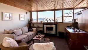 A cozy living room with a wooden ceiling, a fireplace, a sofa, a coffee table, and a view of houses through large windows. - Sugarloaf Inn (Kingfield)