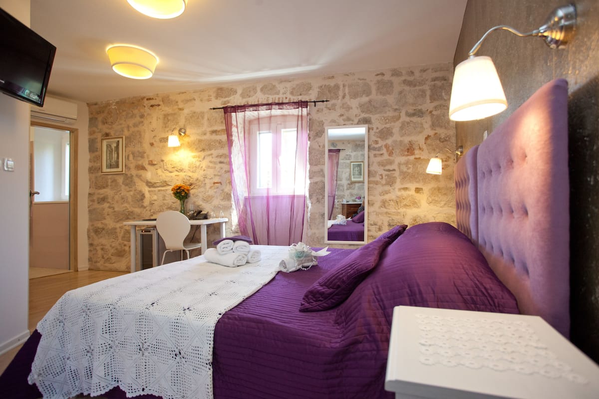 Un dormitorio con pared de piedra, una cama con ropa de cama púrpura, una mesita de noche blanca, un televisor montado en la pared y una ventana con cortinas.
