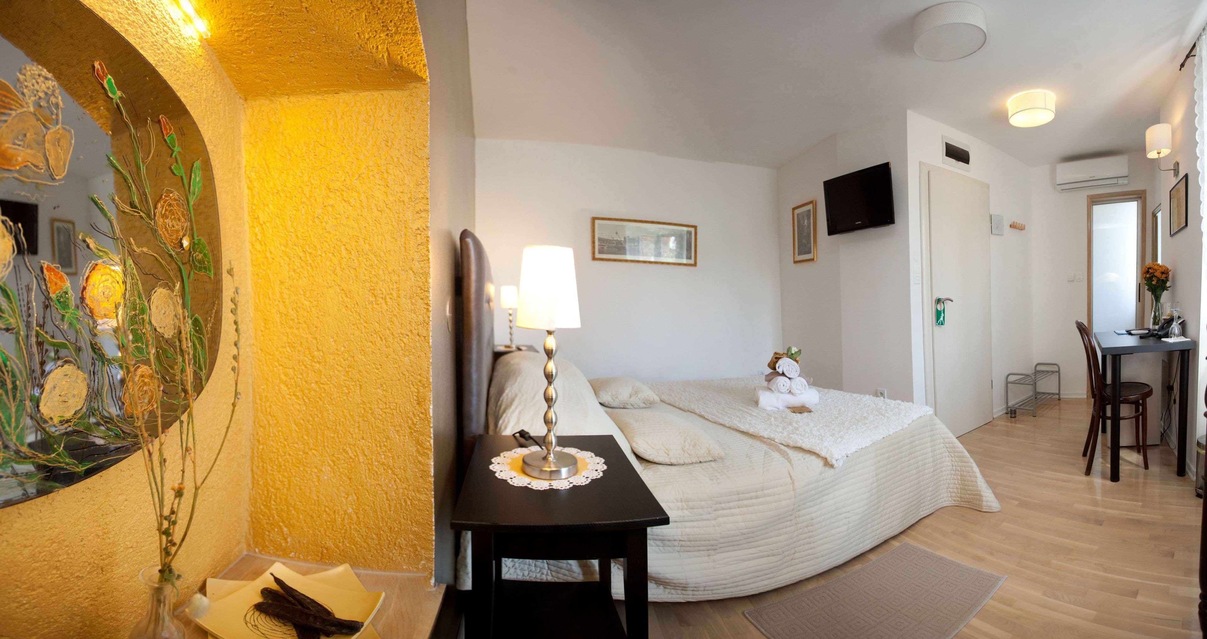 A hotel room with a bed, bedside table, lamp, artwork, and a TV.