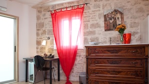 Une pièce avec un mur en pierre, une commode en bois, un bureau avec une chaise, un rideau rouge et un vase rempli de fleurs.