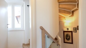 Un escalier avec des marches en bois et une rampe en verre, menant à un palier où se trouvent un portrait encadré et une lampe fixée au mur.