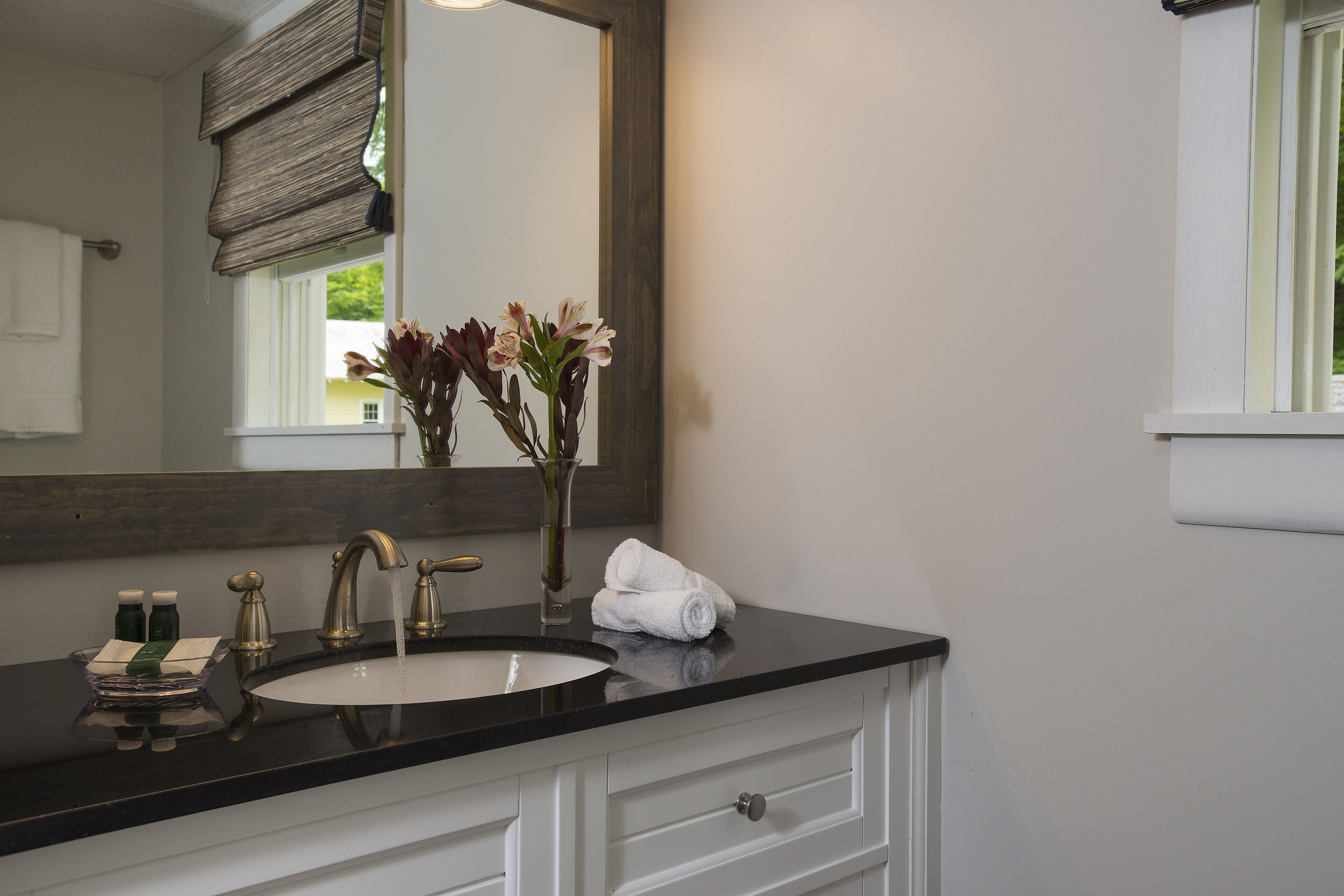 Une salle de bain avec un comptoir noir, un lavabo blanc et un miroir au-dessus. On y trouve une serviette et un vase avec des fleurs sur le comptoir.