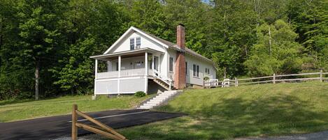 A white two-story house with a front porch and a brick chimney, surrounded by lush green trees.