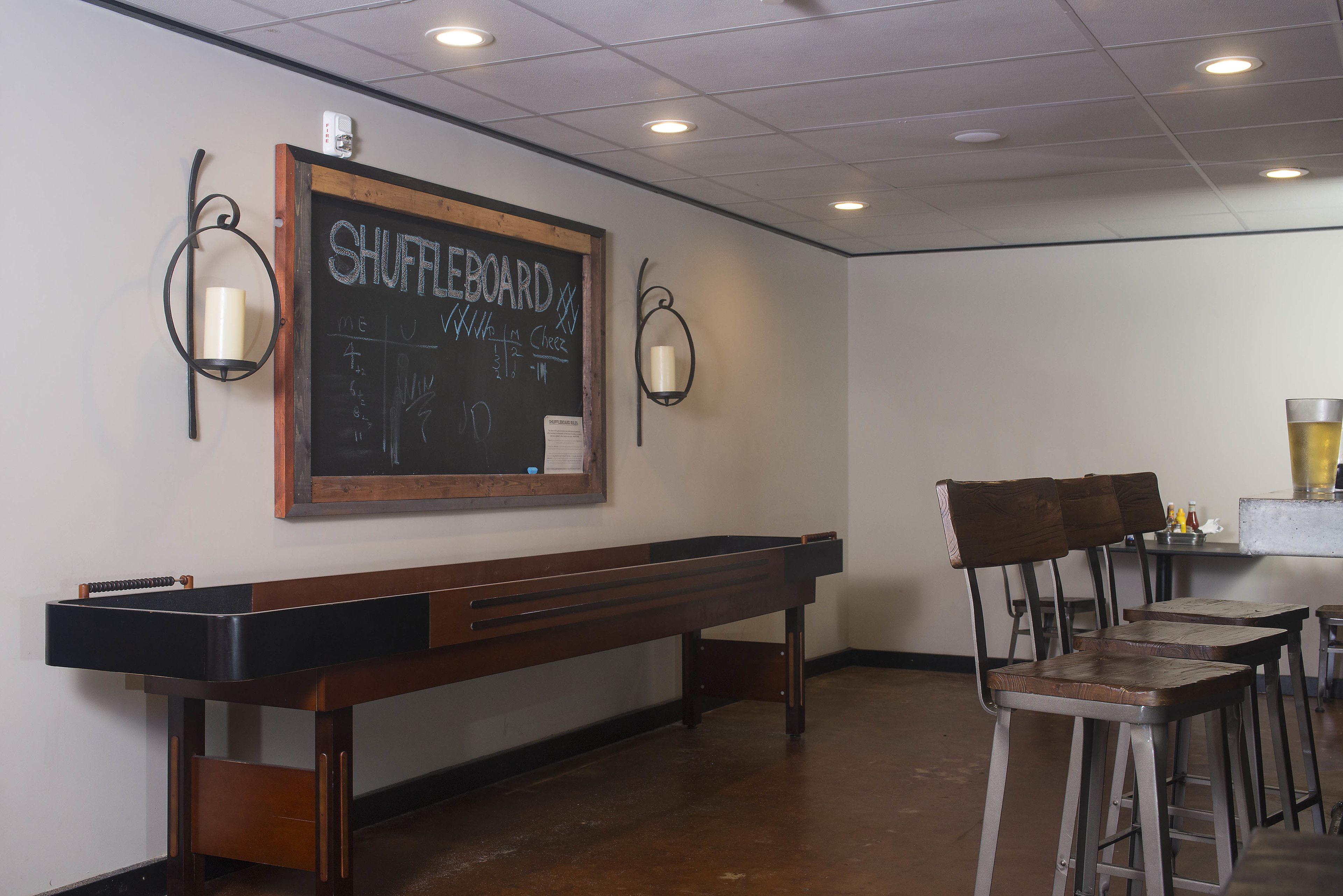 A room with a shuffleboard table, bar stools, and a chalkboard with 'SHUFFLEBOARD' written on it.