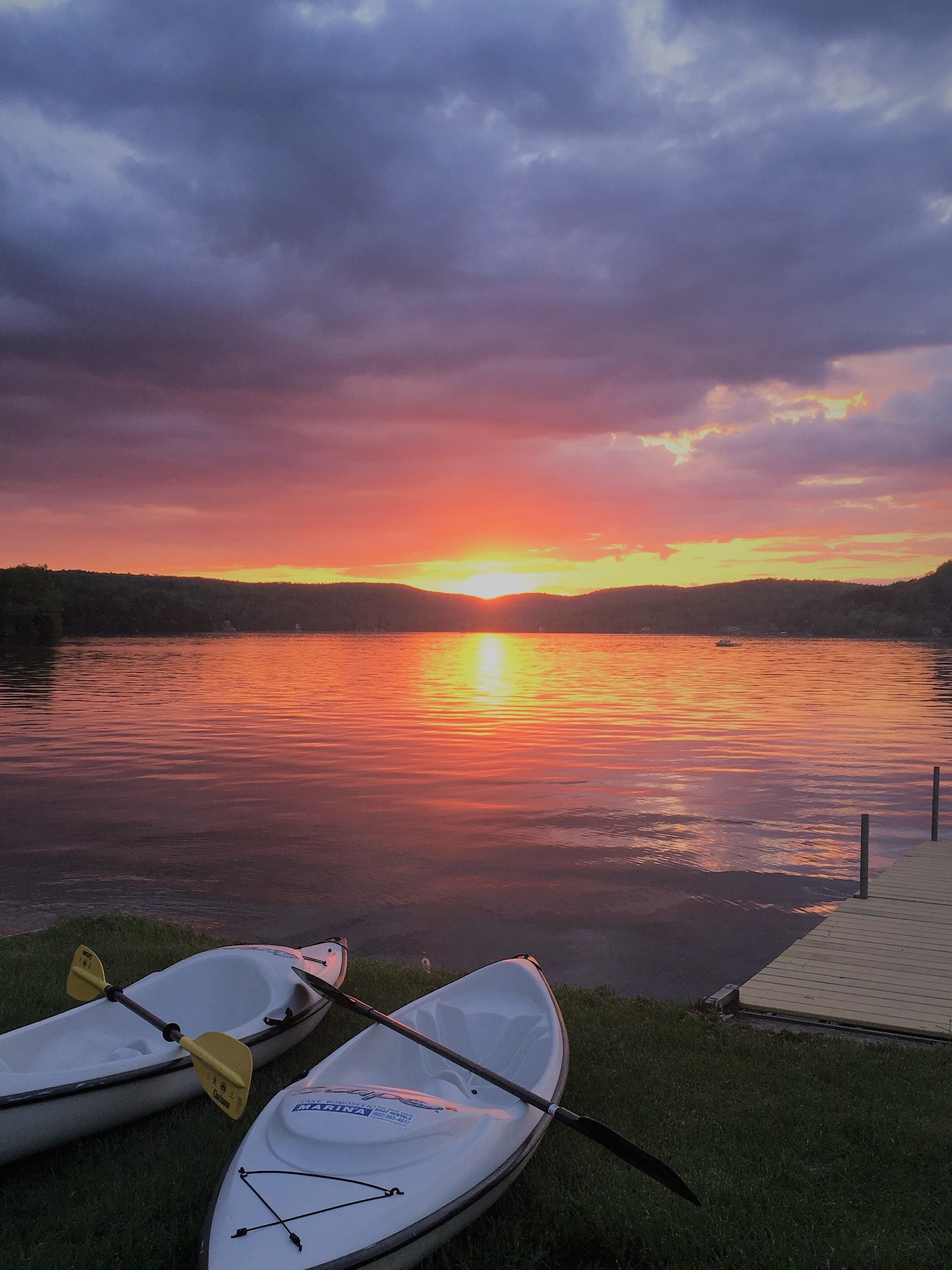 Two kayaks on a grassy shore with a sunset over a calm lake.