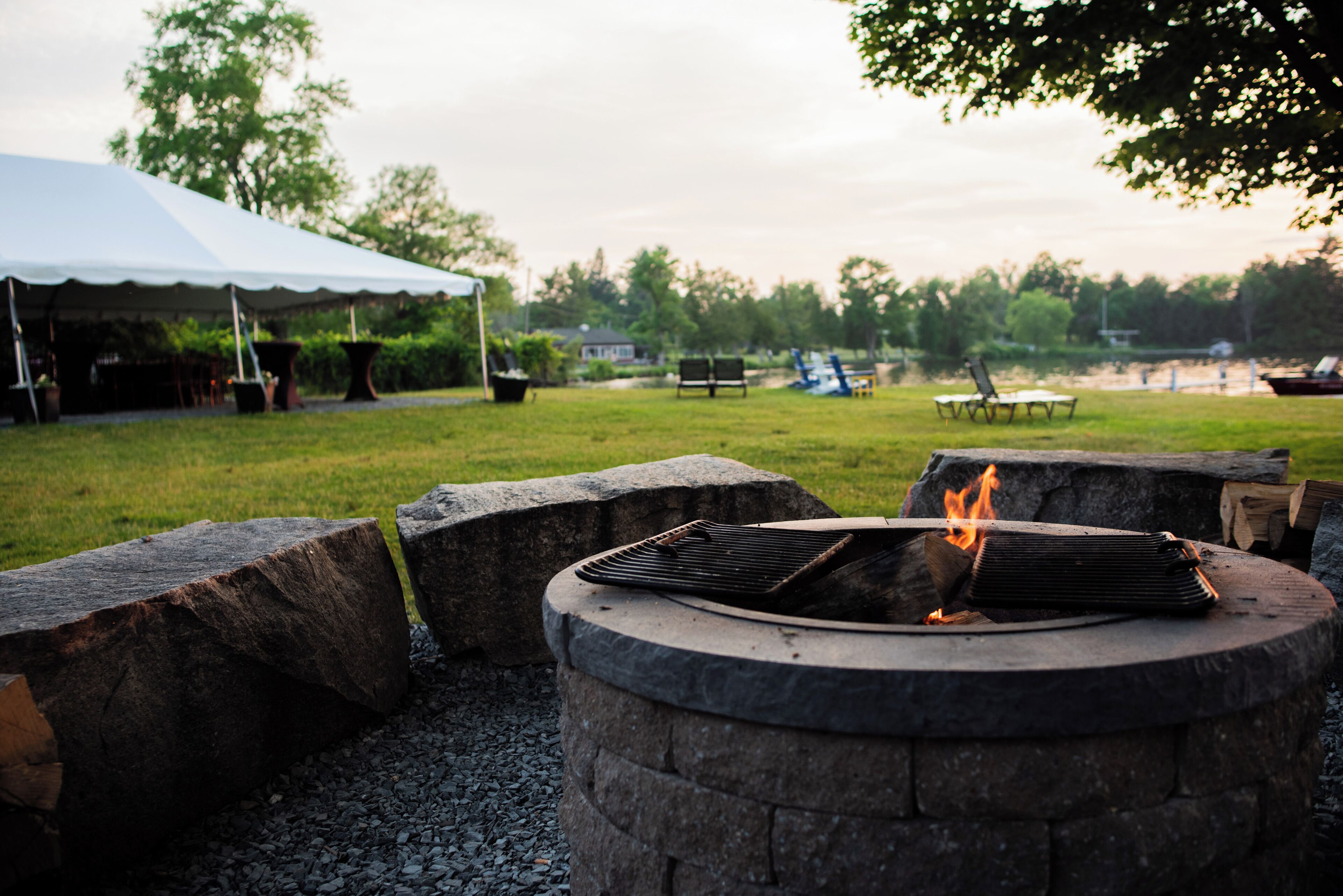 A campfire with a grill, surrounded by stone seating and a tent in a grassy outdoor area.