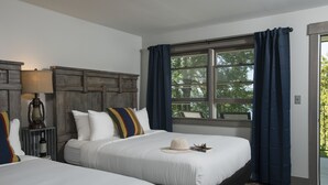 Habitación de hotel con dos camas, una mesita de noche con lámpara, una ventana con cortinas y vistas a árboles.