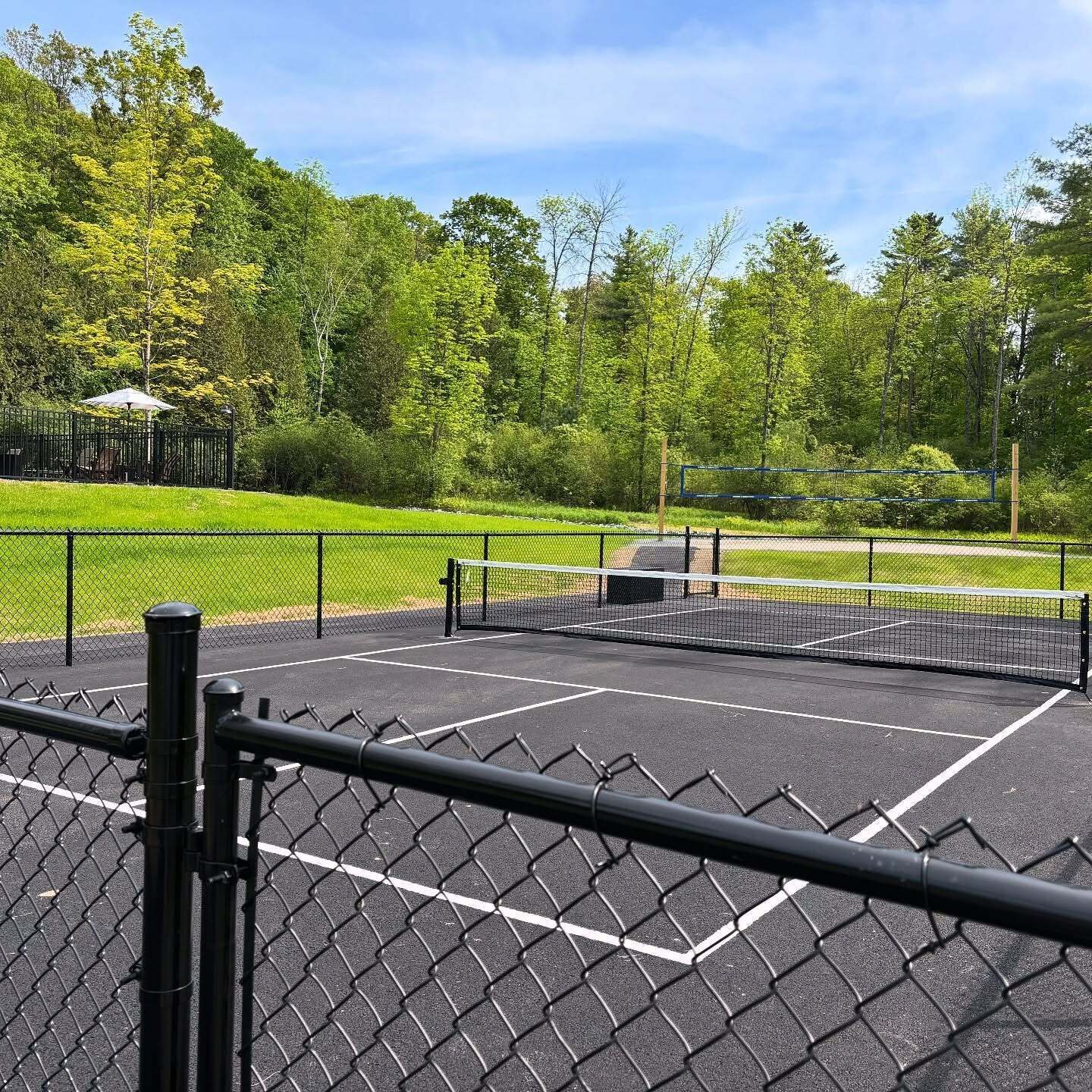 A tennis court with a net, surrounded by a chain-link fence, and a gazebo in the background.