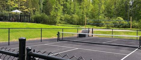 A tennis court with a net, surrounded by a chain-link fence, and a gazebo in the background.