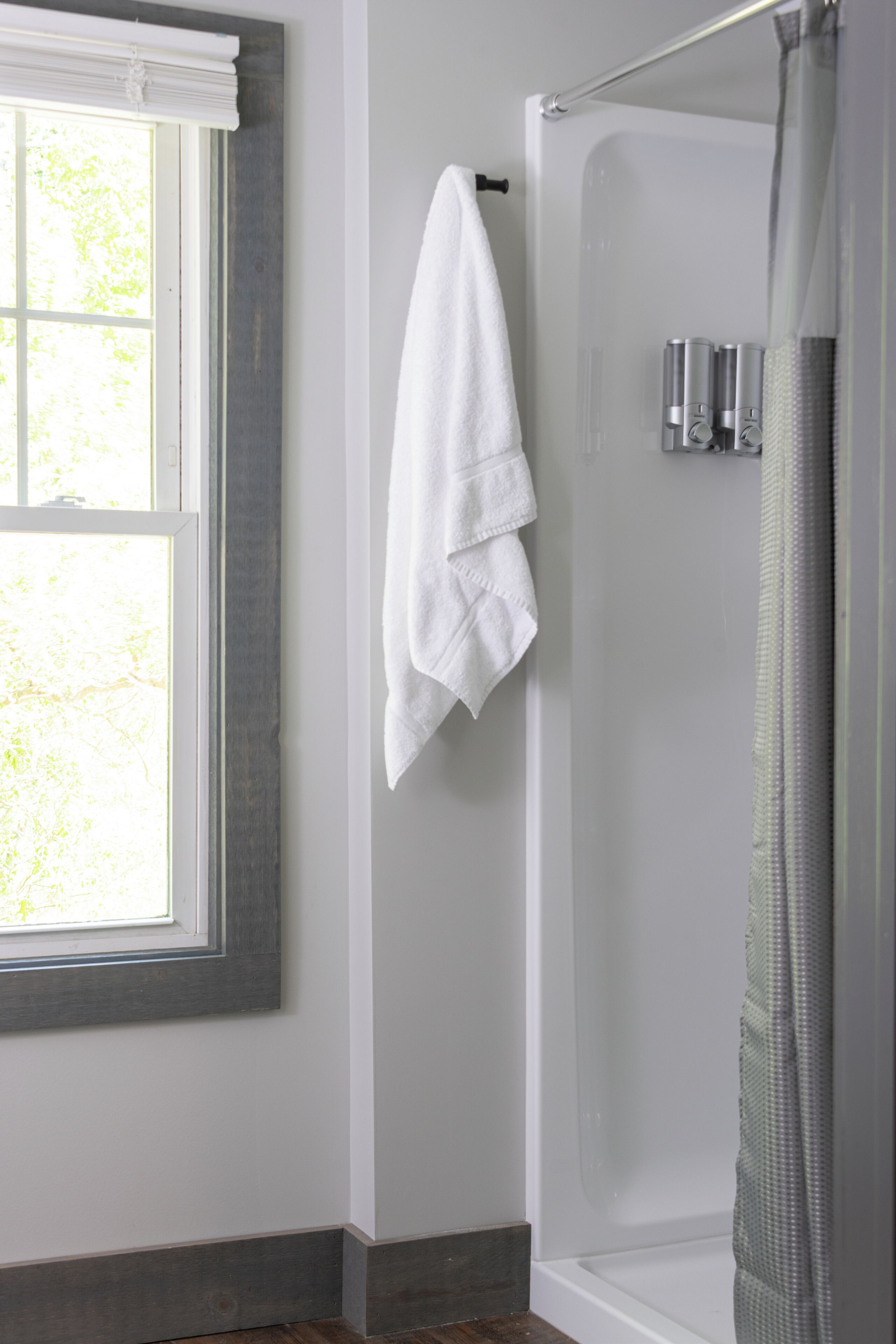 A bathroom with a shower, a towel hanging on the wall, and a window with blinds.