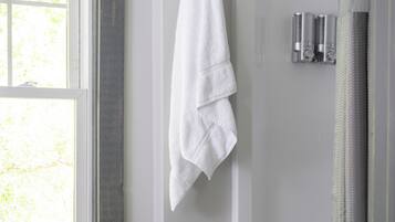 A bathroom with a shower, a towel hanging on the wall, and a window with blinds.