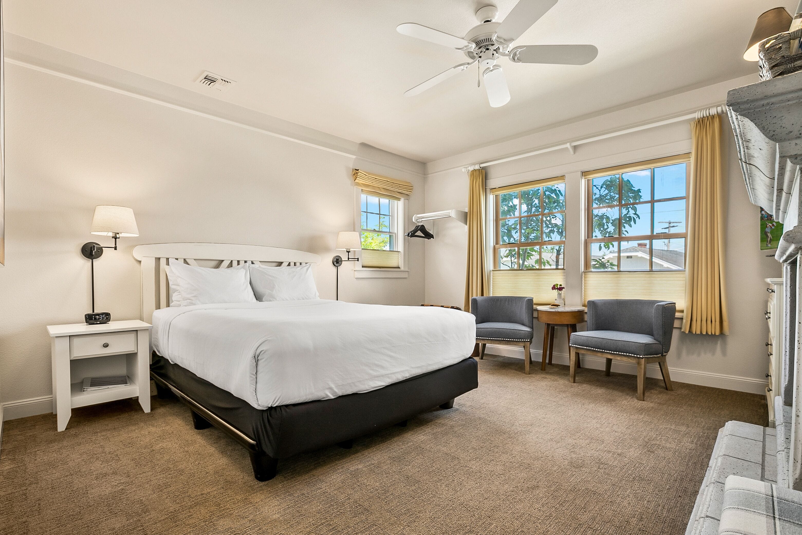 Une chambre d’hôtel avec un grand lit, une table de chevet, une chaise, une petite table et un ventilateur de plafond.