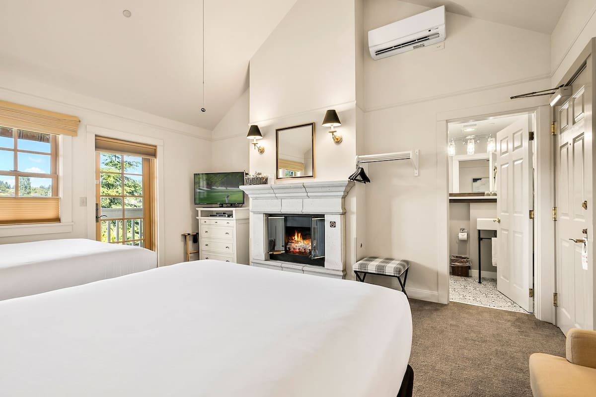 Habitación de hotel con dos camas, chimenea, televisor y baño visible a través de una puerta abierta.
