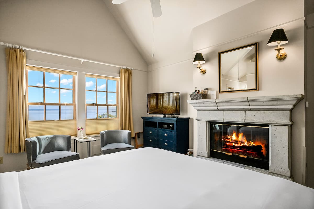Habitación de hotel con una cama grande, dos sillones, una mesita, televisión, chimenea y vistas al mar desde las ventanas.