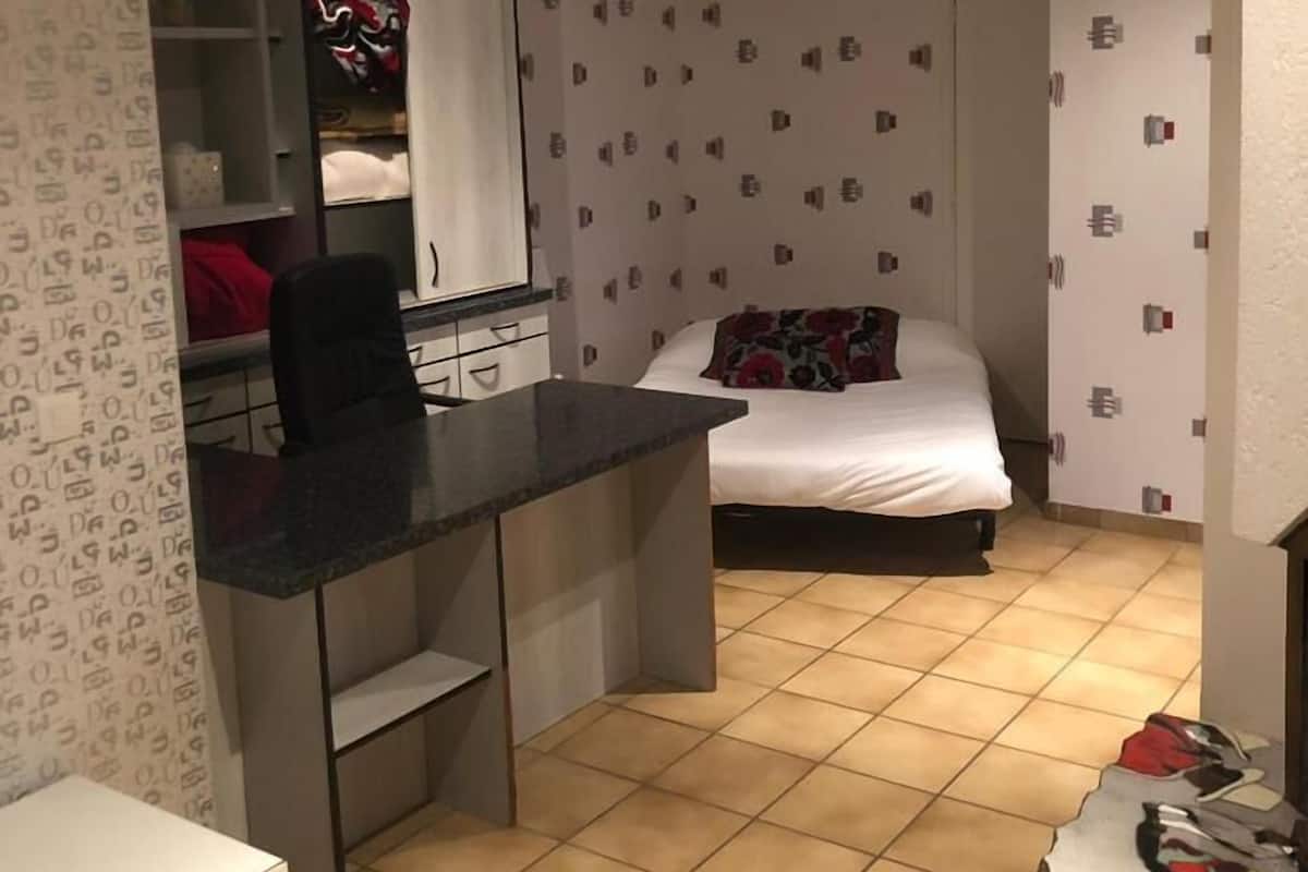 Une petite chambre bien éclairée, avec un lit, un bureau et une armoire.