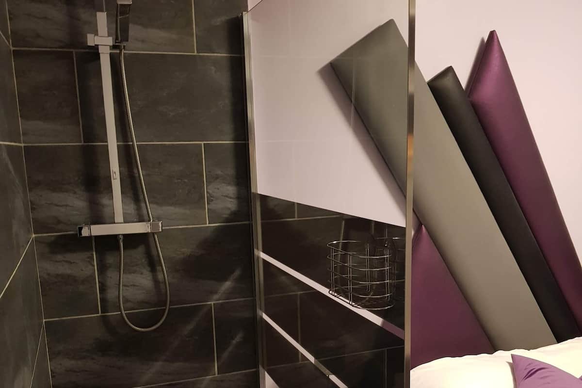 Une salle de bain moderne avec une douche à parois de verre, des murs recouverts de carreaux foncés et un lit avec une literie blanche et un oreiller violet.