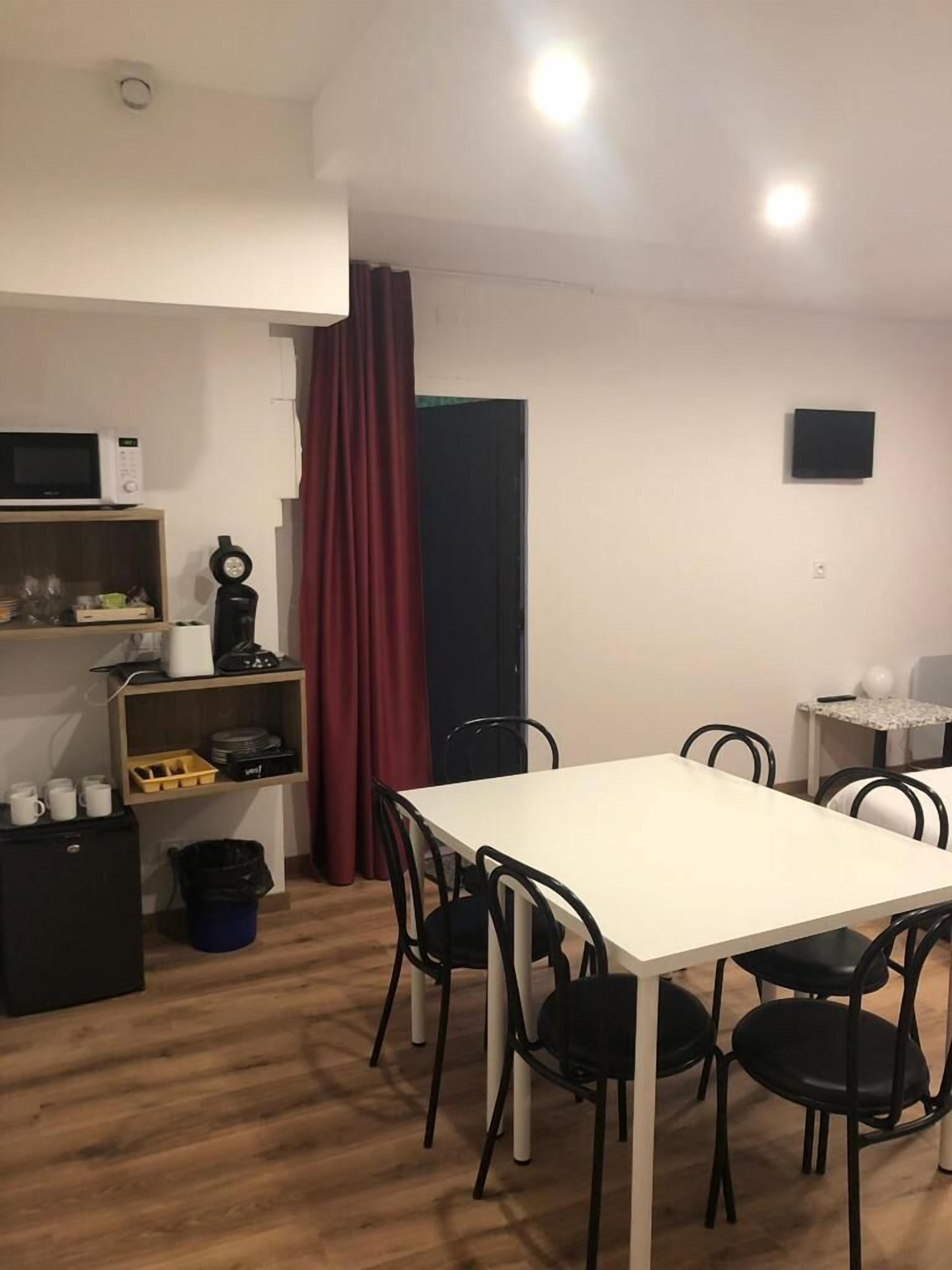 A compact room with a white table and four chairs, a microwave, a coffee maker, and a small shelf with dishes and cups.