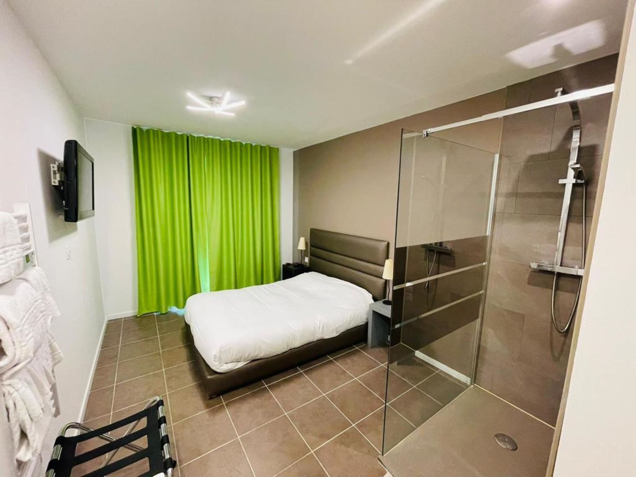 A hotel room with a bed, a shower area, and a television on the wall.