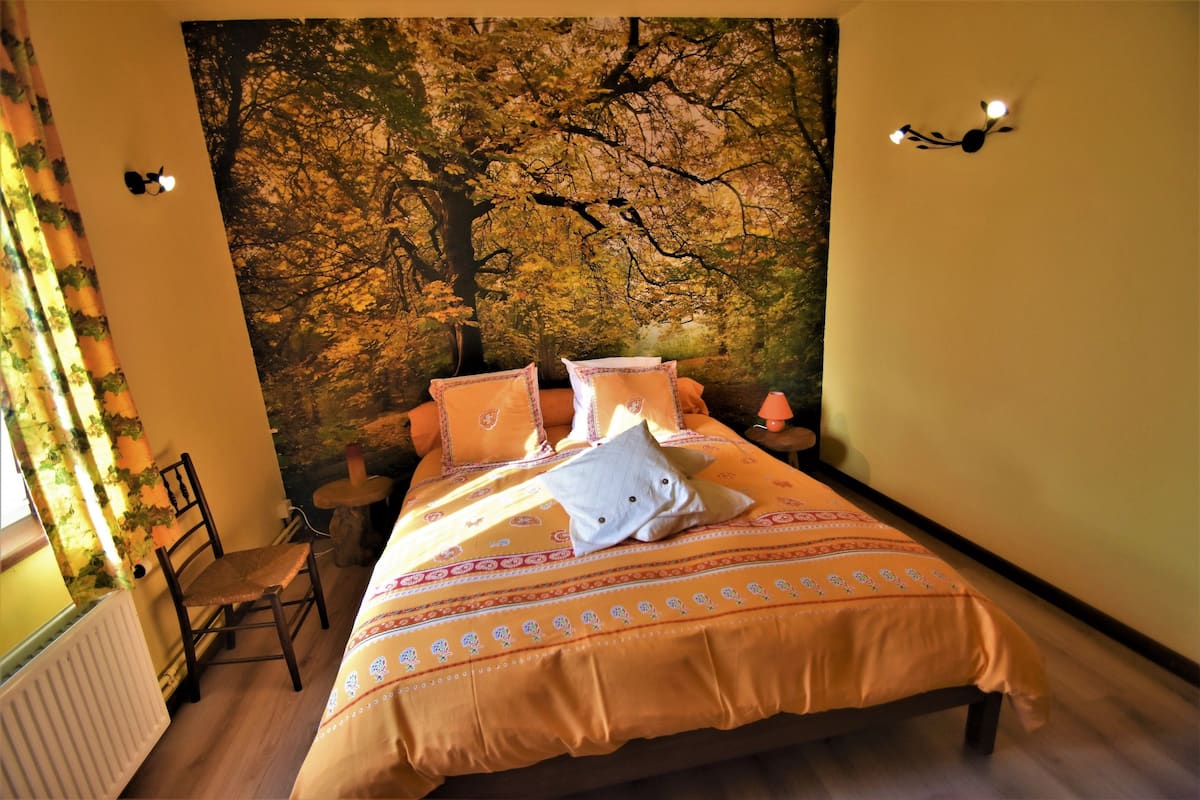 Ein Schlafzimmer mit einer großen Wandmalerei von einem Baum, einem Bett mit orangefarbener Bettwäsche, einem Sessel und einem kleinen Tisch.