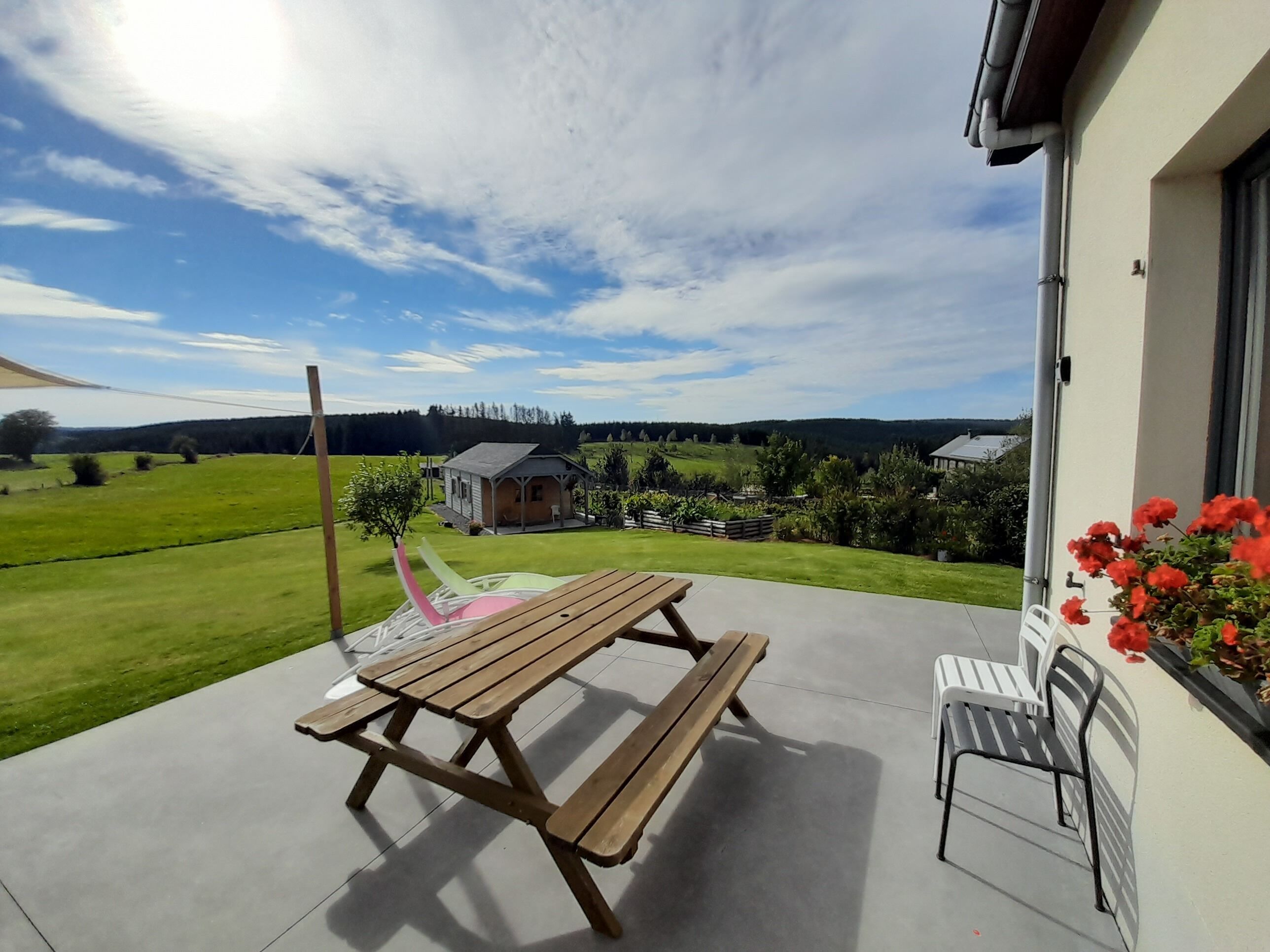 Eine Terrasse mit Holztisch und -stühlen, Blick auf ein grünes Feld und ein kleines Gebäude in der Ferne.